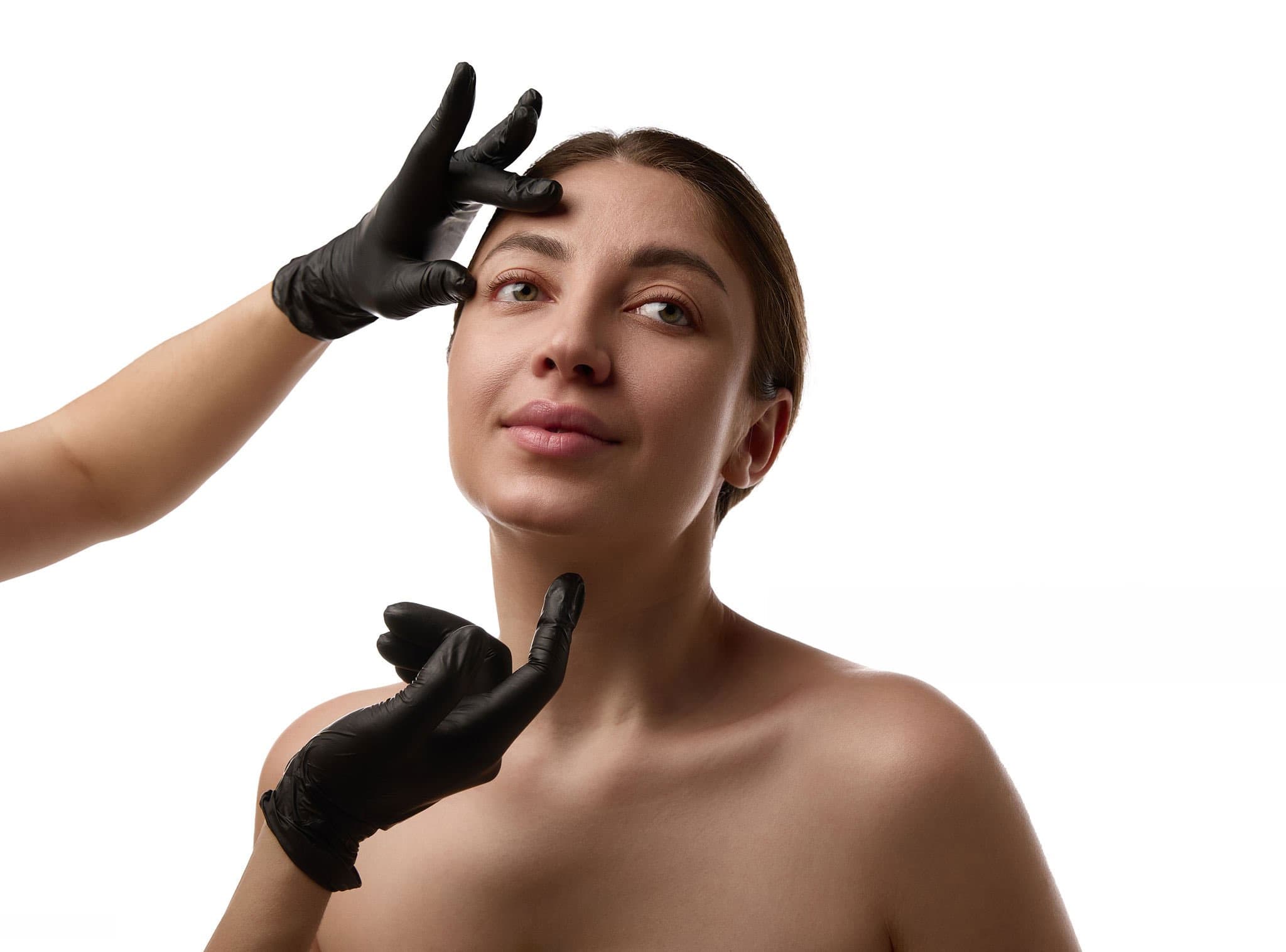 Gloved hands frame a woman's forehead and chin while assessing facial proportion