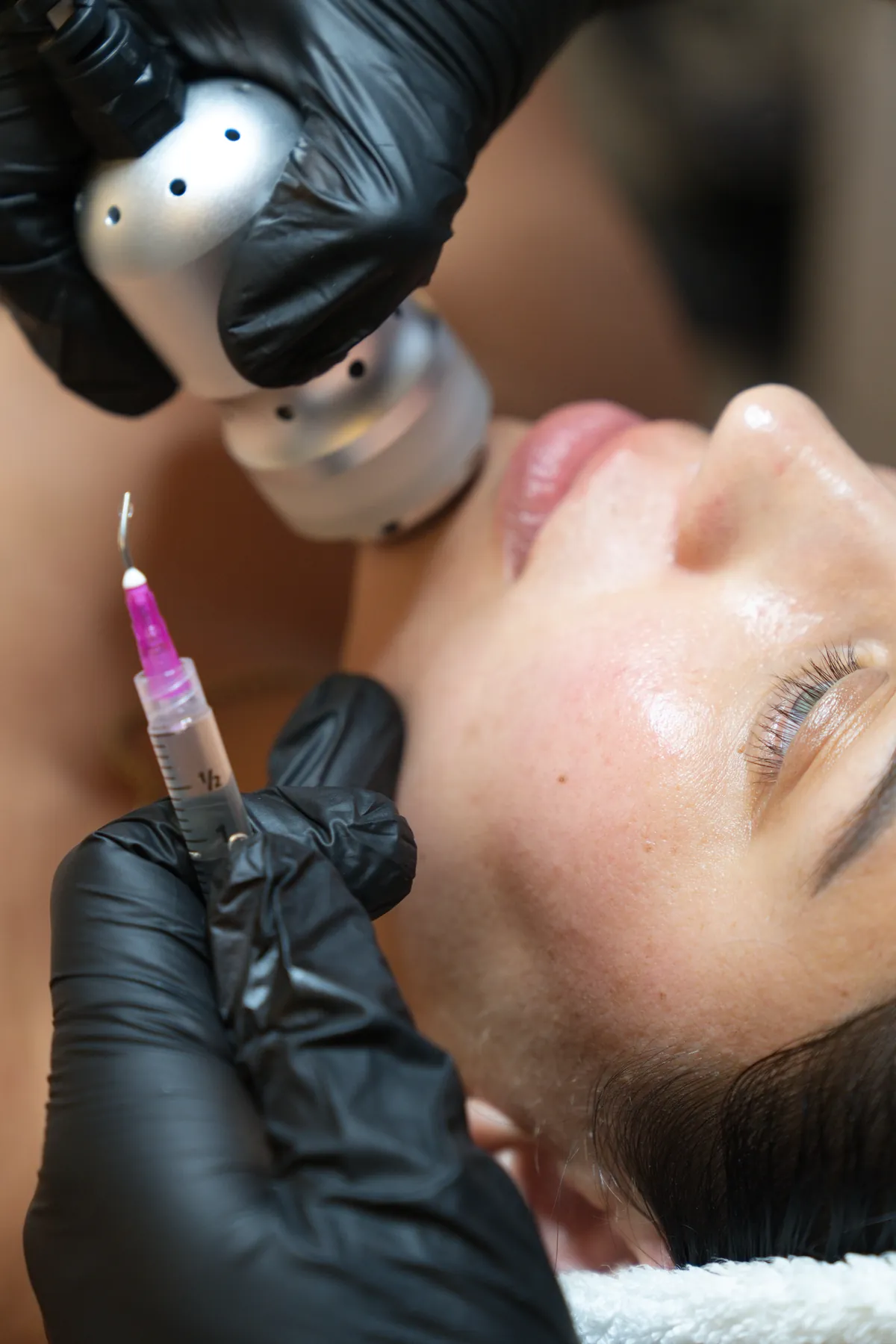 Gloved hands hold a pink-tipped syringe near a woman's face while a silver handheld device rests along her jawline.