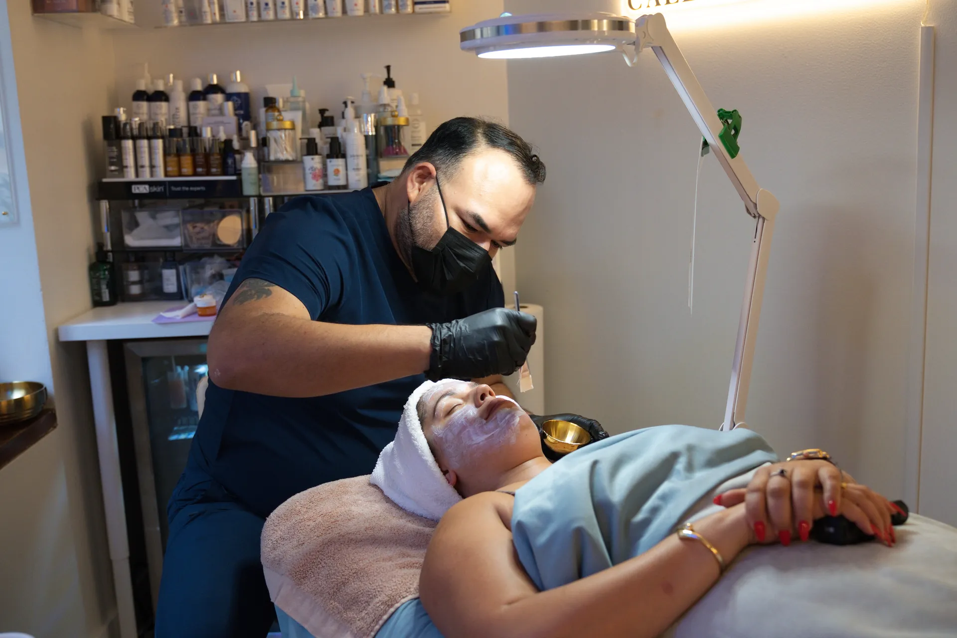 A masked provider in black gloves performs a facial on a woman lying under a blue blanket, with a magnifying lamp and skincare products in the treatment room.