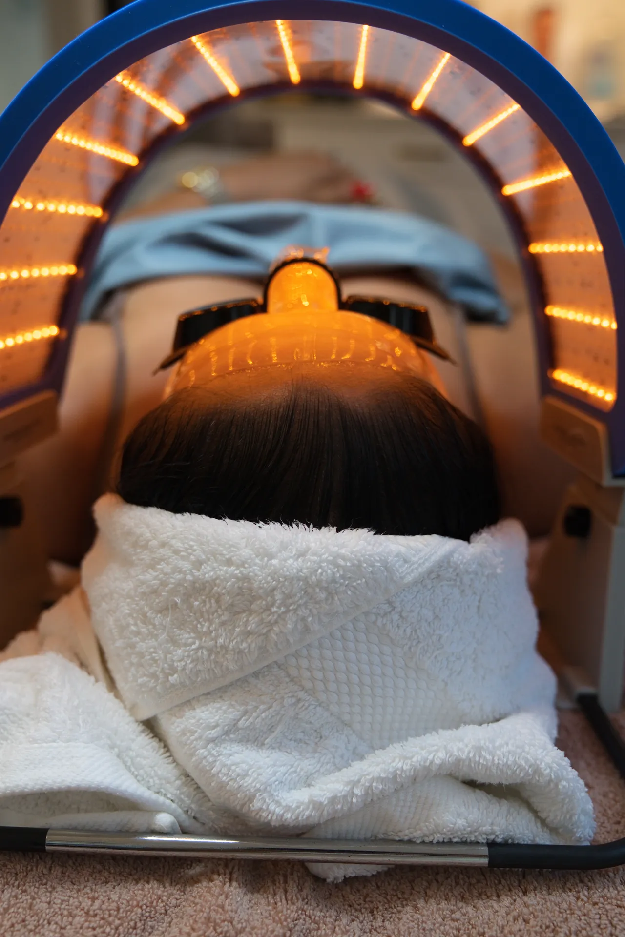 A woman lies under a curved LED light dome glowing orange-red, with a white towel wrapped around her shoulders.