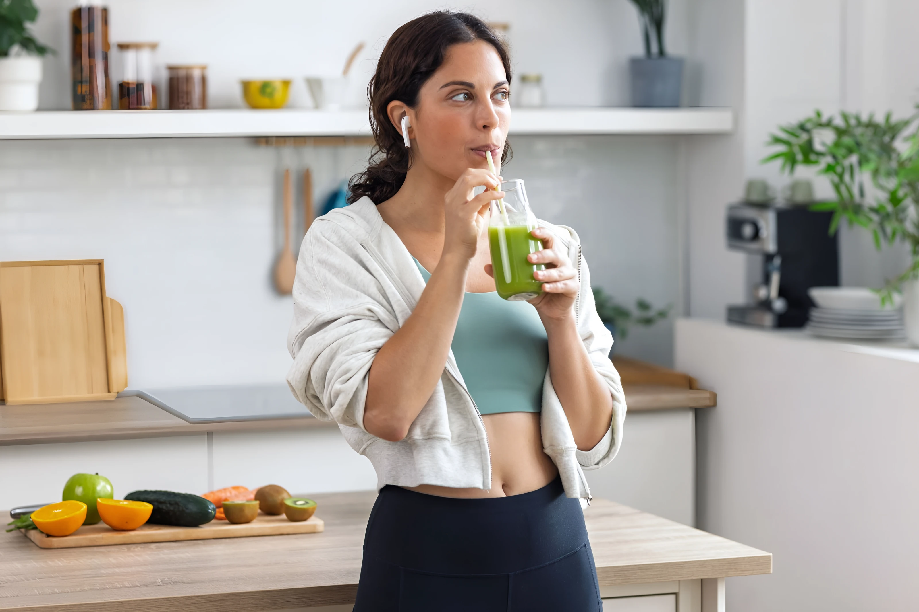Mujer en la cocina bebiendo un batido verde, con fruta fresca en la encimera
