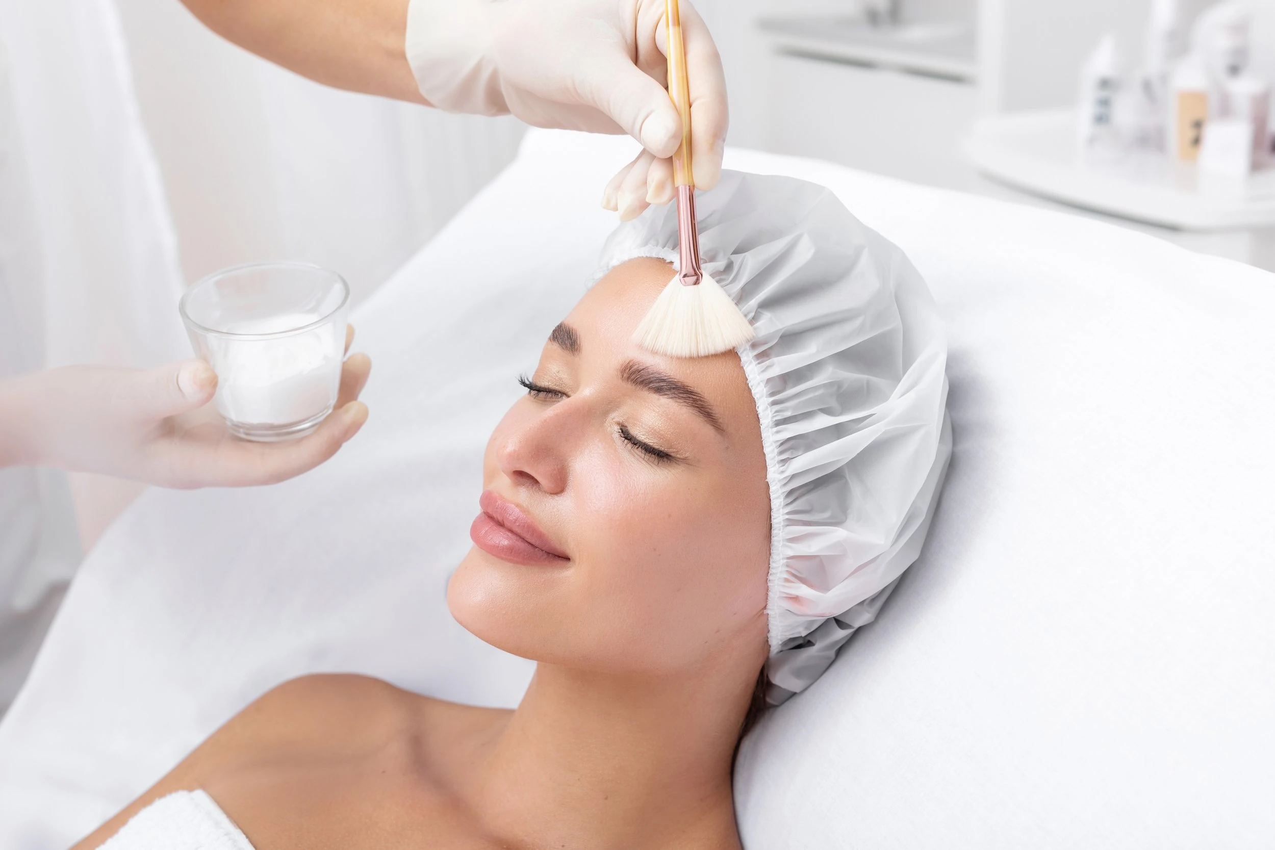 Gloved hands brushing a peel onto the forehead of a woman wearing a hair cap