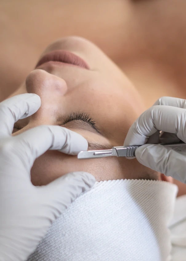 Gloved hands using a dermaplaning blade along a woman’s cheek